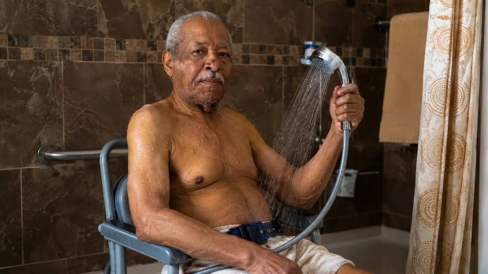 Elderly man confidently showering seated, handheld showerhead in hand, calm expression