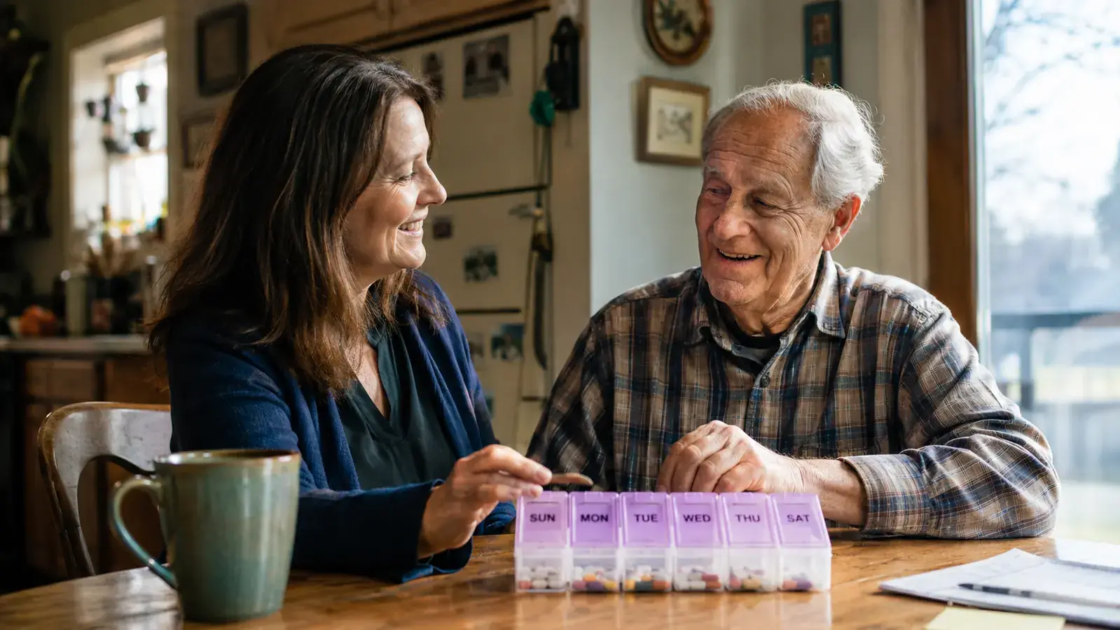 Adult daughter and elderly father sitting together at a kitchen table, smiling, a pill organizer visible between them